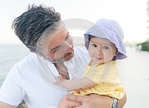 outdoors lifestyle portrait of father and little daughter - man holding his adorable and happy baby girl in front of the see