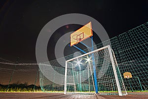 Outdoors mini football and basketball court with ball gate and basket surrounded with high protective fence brightly illuminated