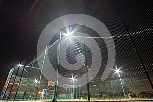 Outdoors mini football and basketball court with ball gate and basket surrounded with high protective fence brightly illuminated