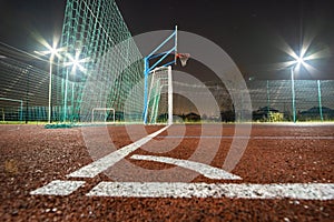 Outdoors mini football and basketball court with ball gate and basket surrounded with high protective fence brightly illuminated