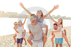 Outdoors photo of happy boyfriends piggybacking their girlfriends at sunset on beach