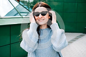 Outdoors portrait of beautiful young brunette girl smiling, close up. Teenager hipster girl with sunglasses wearing trendy outfit