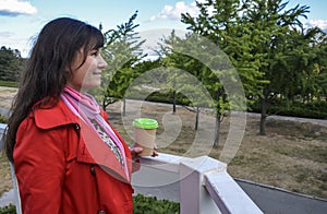 Outdoors portrait of cheerful beautiful brunette woman in red coat holding disposable coffee cup drinking coffee, and smiling