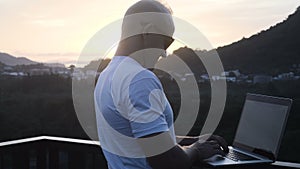 Outdoors portrait of handsome young man working on a laptop computer while standing on a rooftop over beautiful sunrise