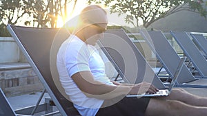 Outdoors portrait of handsome young man working on a laptop while sitting on sunbed on a rooftop