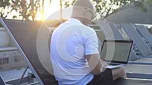 Outdoors portrait of handsome young man working on a laptop while sitting on sunbed on a rooftop