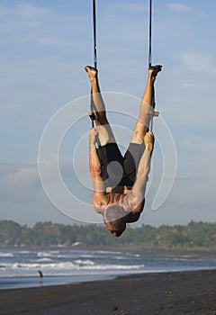 Outdoors portrait of  man practicing aero yoga workout at the beach hanging from rope swing above the sea training body balance