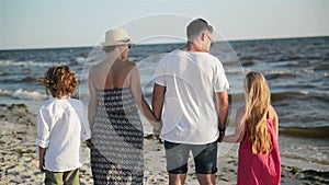 Outdoors Portrait of Mother, Father, Son and Daughter Holding Hands on the Beach During Sunny Windy Day near the Sea.