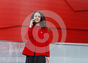 Outdoors portrait of a smiling brunette young woman, wear in red, talking on mobile phone, over building background.