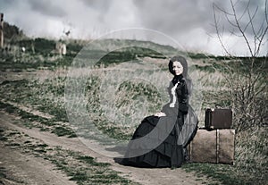 Outdoors portrait of a victorian lady in black sitting alone on the road with her luggage