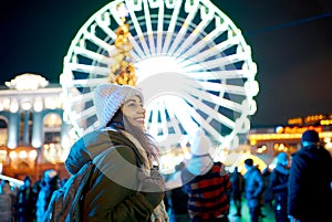 Outdoors portrait winter woman at everning festive Christmas fair with ferris wheel on background.