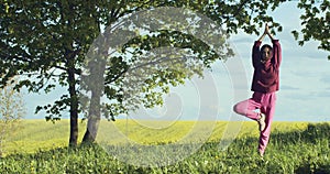 Outdoors portrait of woman standing in tree position vrikshasana on nature background. Sportive girl in hood doing yoga