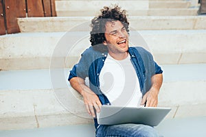 Outdoors shot of cheerful male with curly hair using laptop for chatting online with friends, connected to free wireless on the
