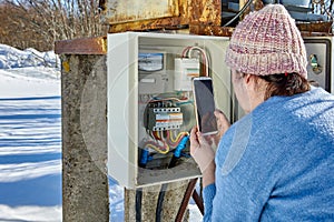 Outdoors switchboard with electric meter, woman takes readings using smartphone in winter.