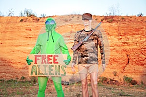 Outdoors in the Utah desert two men one military mannequin second alien against the background of the mountains holding a sign in