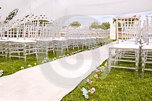Outdoors, wedding aisle with a white carpet, flowers, a white wedding tent and glass chairs