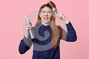 Outraged Caucasian woman has angry expression, combs unmanageable hair, dissatisfied with new conditioner, holds hairspray, fails