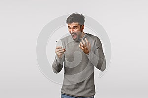 Outraged and furious pissed, tensed bearded man in grey sweater, holding smartphone, cursing, swearing at mobile display