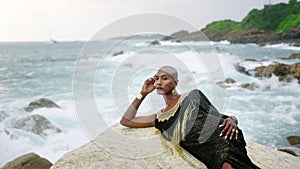 Outrageous LGBTQ bipoc person in luxury dress, boutique brass jewelry poses on cliff top, against wavy ocean. Gender