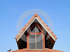 Outside of old house attic with glass window red wood frame and bird perched on orange roof tiles