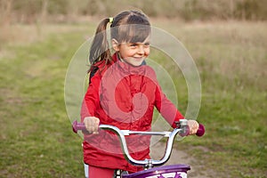 Outside picture of cheerful joyful little girl with pony tails, riding bicycle alone, having fun, breathing fresh air, being fond