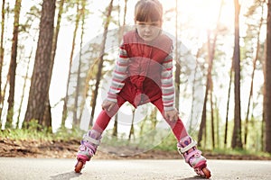 Outside picture of energetic serious little girl being in forest, having roller blades on legs, enjoying active lifestyle, having