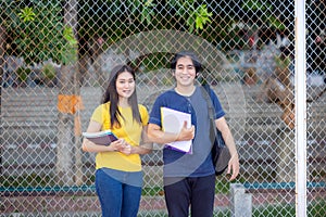Outside of school, a happy young couple of students stands along a fence, studying a book