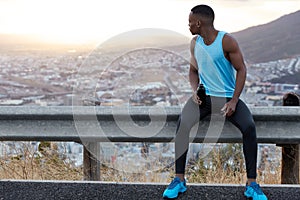 Outside shot of dark skinned man focused backwards, wears casual sportsclothes, holds bottle with cold water, takes