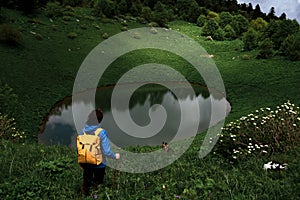 Oval mountain lake among alpine meadows. Female hiker looks at lake. Girl travels in summer through mountains of Caucasus with her