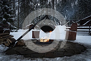 Over fire hangs a pot in which to cook food. On a hook on a tripod, steam comes out of the pan. Winter Camping outdoor cooking