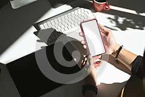 Over head shot of woman holding mobile phone mockup while sitting on her workspace.