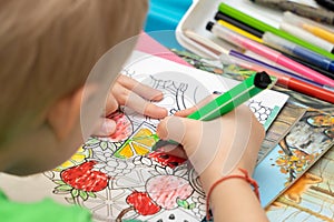 Over the shoulder: a boy learns to color a coloring book with fruits and vegetables with a green felt-tip pen. Development of