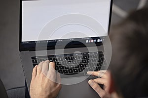 Over the shoulder of a man typing on a laptop with a blank white screen, with computer on his laps and fingers on his keyboard