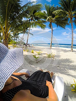 Over shoulder POV: woman in hat and dress on sun lounger on vacation at idyllic tropical beach with palm trees at Lefaga, Upolu I photo