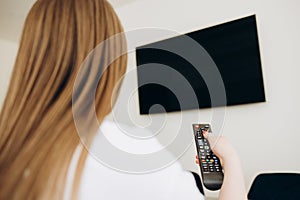 Over the shoulder view of young Asian girl with long black hair sitting on sofa holding tv remote and surfing programs