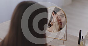 Over shoulder view on young middle eastern lady looking at mirror and applying moisturizing cream on her cheeks