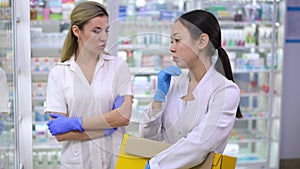 Overburdened Asian young pharmacy assistant with box standing at shelves with angry Caucasian manager arguing at