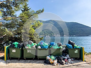 Overflowing garbage cans stand in a fence on the seashore near a tree