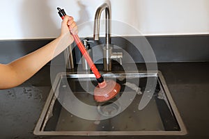 Overflowing kitchen sink, clogged drain. Hand holding plunger.