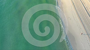 overhead aerial shot of a smooth sanding beach with green ocean water and waves rolling into the beach, birds and a woman walking