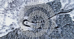 Overhead aerial top view over car travelling on hairpin bend turn road in mountain winter snow covered forest.White pine