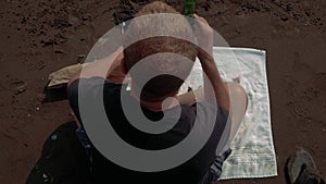 Overhead angle: Jittery man on towel tries to relax at beach with a drink