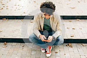 Overhead image of young man sitting on th stairs outdoors typing messages on mobile phone or playing the game. Handsome male with