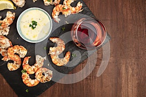 Overhead photo of plate of cooked shrimps on a dark rustic background, with a sauce and a glass of wine, with copy space