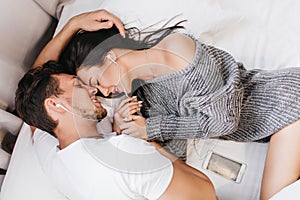 Overhead portrait of brunette man lying in bed with girlfriend. Adorable dark-haired girl chilling with husband in lazy
