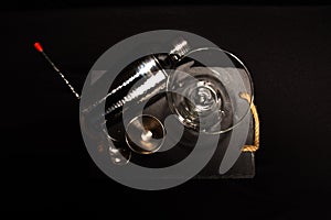 overhead shot with black background of four cocktail instruments including jigger, martini glass, shaker and cocktail spoon