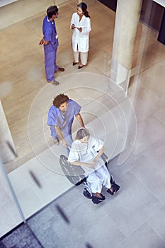 Overhead Shot Of Nurse Wearing Scrubs Pushing Female Patient In Wheelchair Through Hospital Building