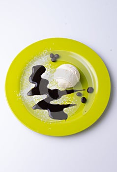 Overhead shot of a scoop of vanilla ice cream on a yellow plate with powdered sugar, chocolate syrup