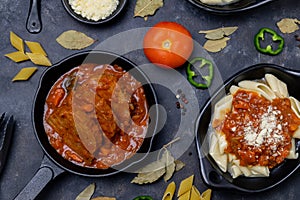 Overhead shot of a a stew dish and pasta, surrounded with ingredients on a black surface