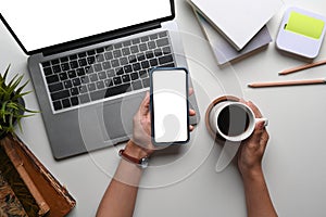 Overhead shot view of woman hand holding coffee cup and using smart phone at her office desk with laptop.
